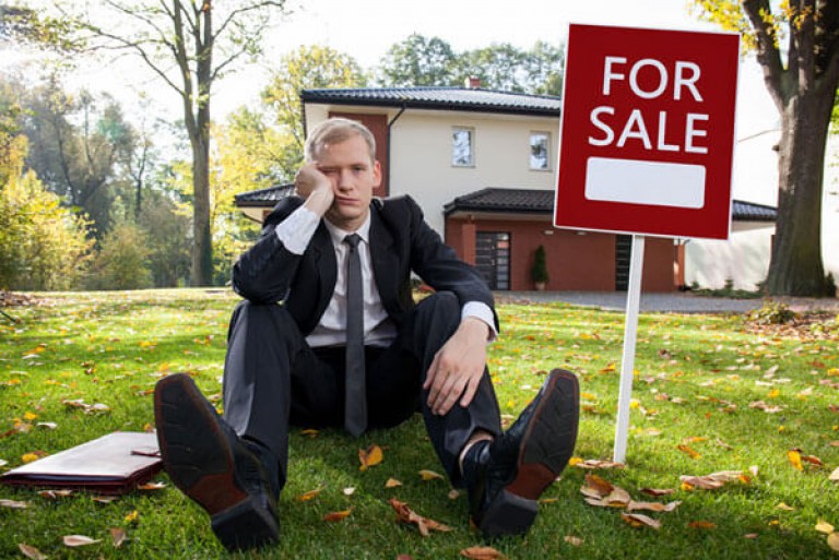 Dejected real estate agent sitting in front of house next to for sale sign - Custodian